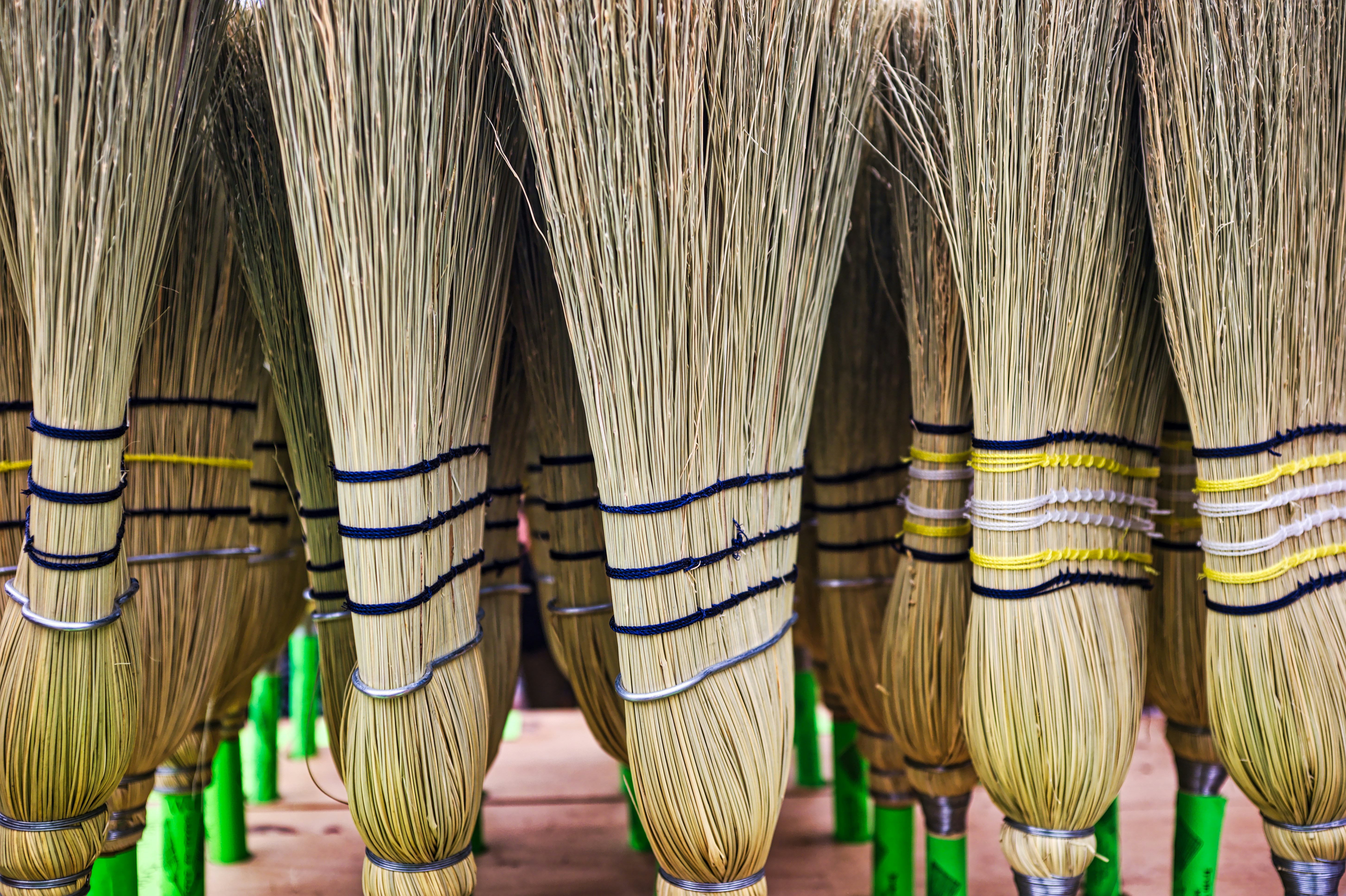 Collection of Handcrafted Grass Brooms Displayed for Sale at Local Market During Sunny Day - A vibrant market showcases a variety of handcrafted grass brooms, neatly arranged for potential buyers. The brooms feature colorful strings wrapped around their handles, highlighting the artisanship involved in their creation. The sun casts a warm glow, enhancing the texture and details of each broom.
